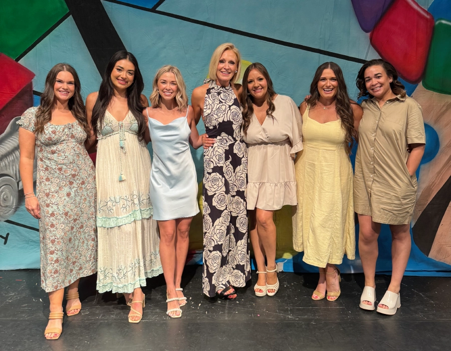 A group of seven women standing together in front of a colorful mural, smiling and posing for a photo. They wear a mix of dresses and casual outfits in light colors.