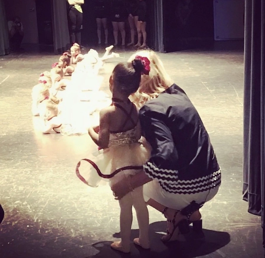 A blonde woman wearing a black jacket and white pants kneels on a dimly lit stage, comforting a young Black girl in a white ballet tutu and leotard, who is looking towards a group of children lying down in white costumes on the stage in front of them.
